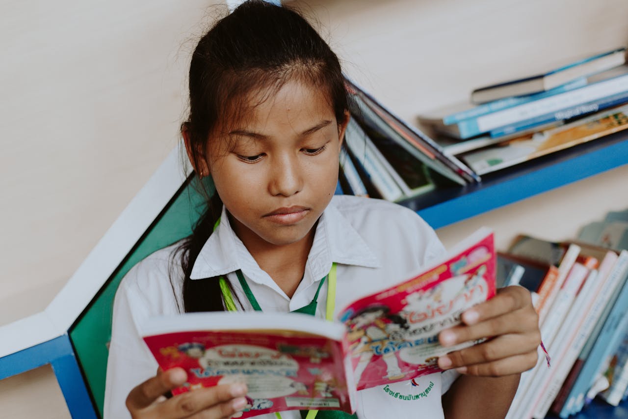 A young girl in a school uniform deeply engaged in reading a book at a library.