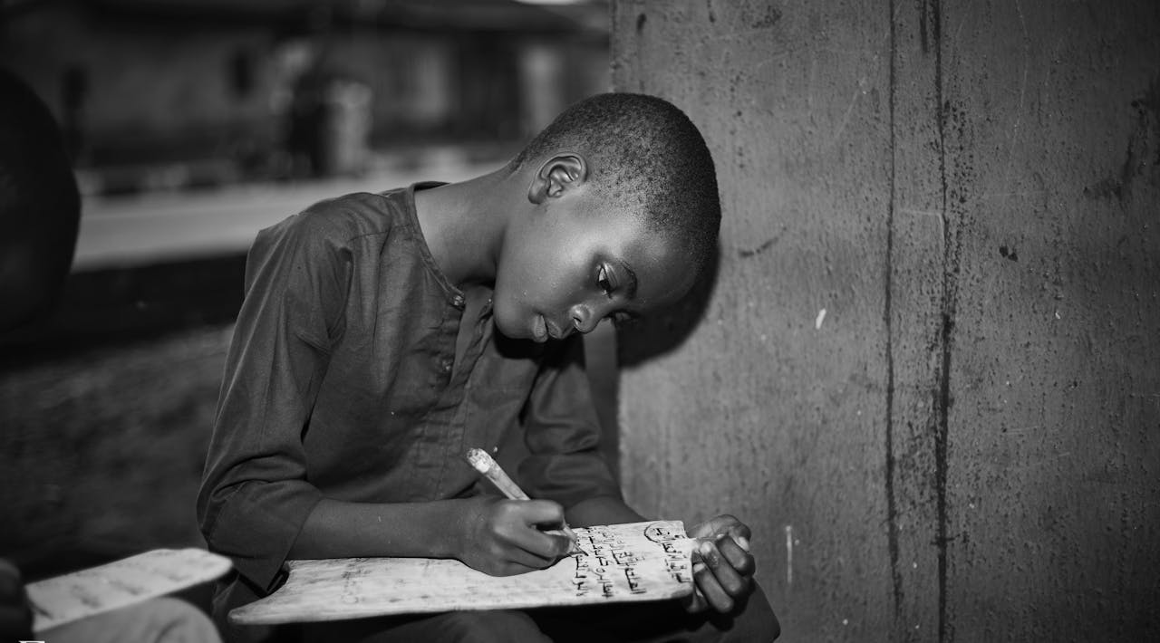 Black and white image of a young boy writing on a slate, focused and determined.