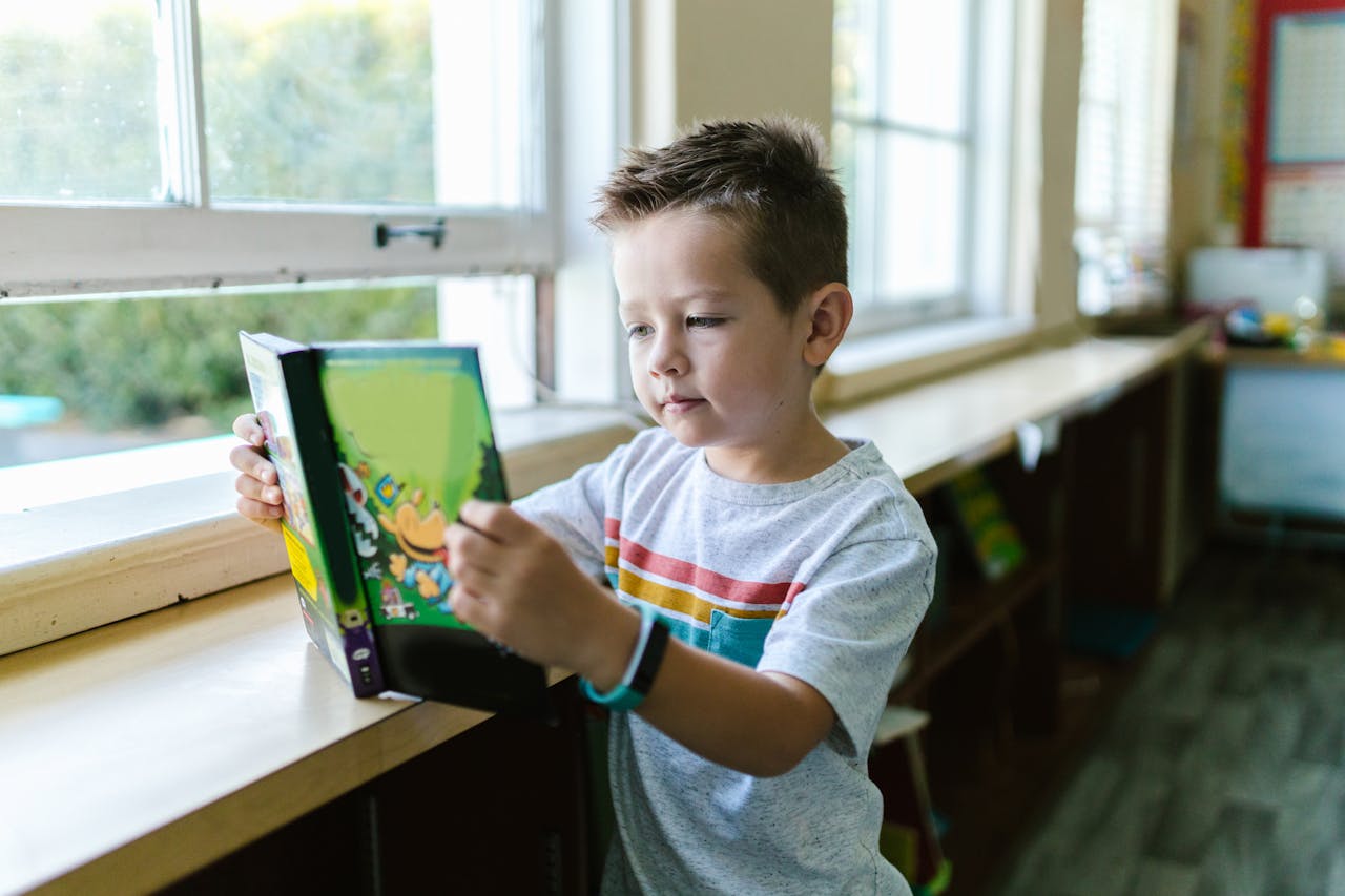 A young boy attentively reads a book in a brightly lit classroom environment.